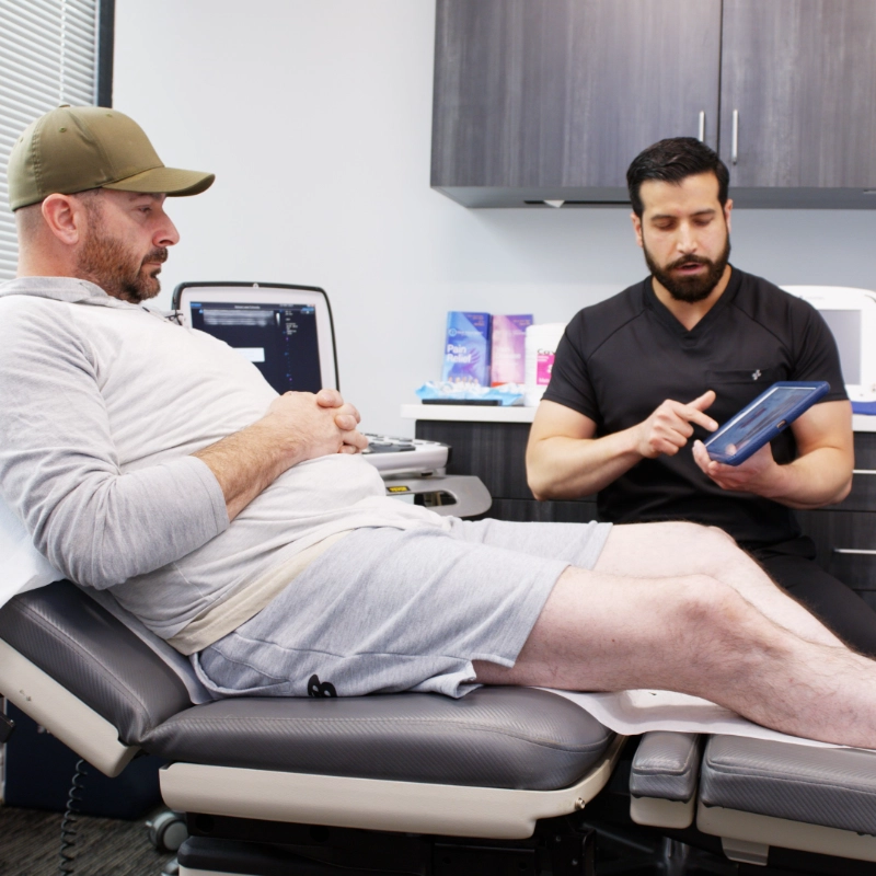 A friendly doctor in a white lab coat shows information about a vein treatment process on a tablet to a seated, smiling patient in a modern medical office setting.