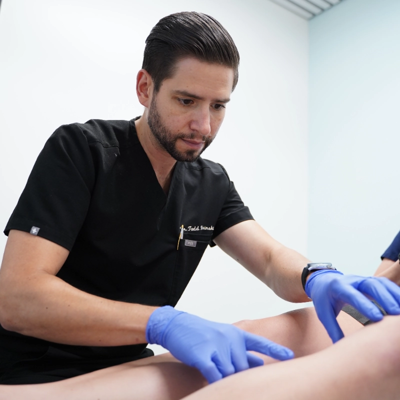 A friendly doctor in a white lab coat shows information about a vein treatment process on a tablet to a seated, smiling patient in a modern medical office setting.