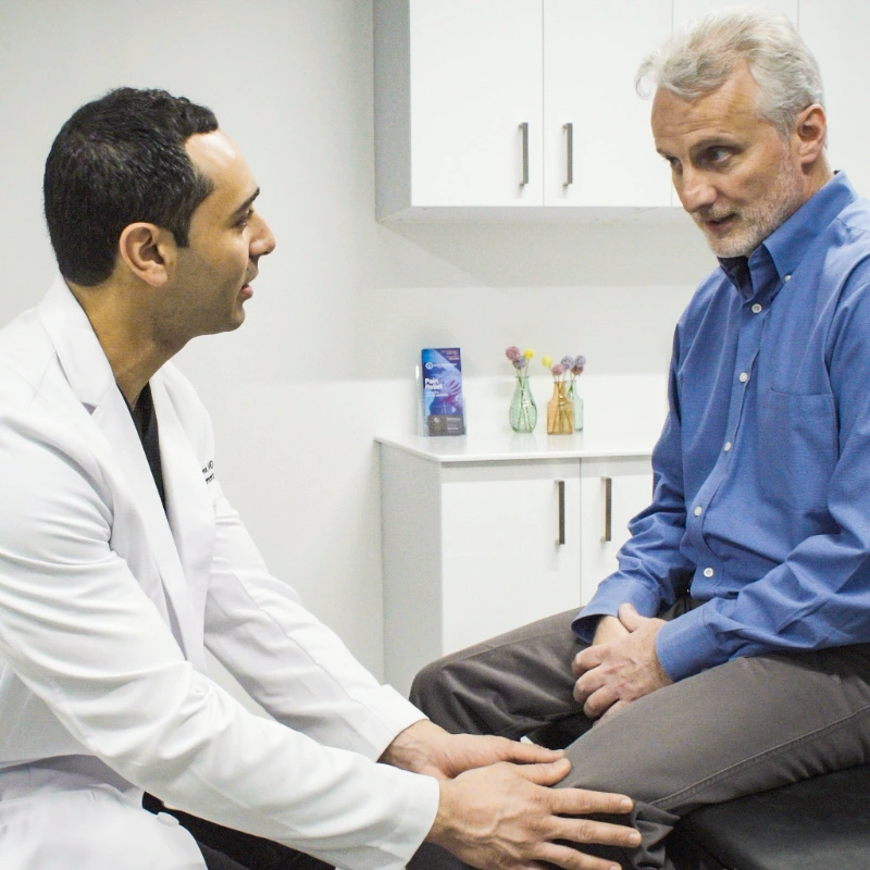 Dr. George Hanna performs a physical examination on an older male patient's knee in a clinical setting. A C-arm fluoroscopy machine and medical monitors are visible in the background, highlighting the interventional pain management environment.