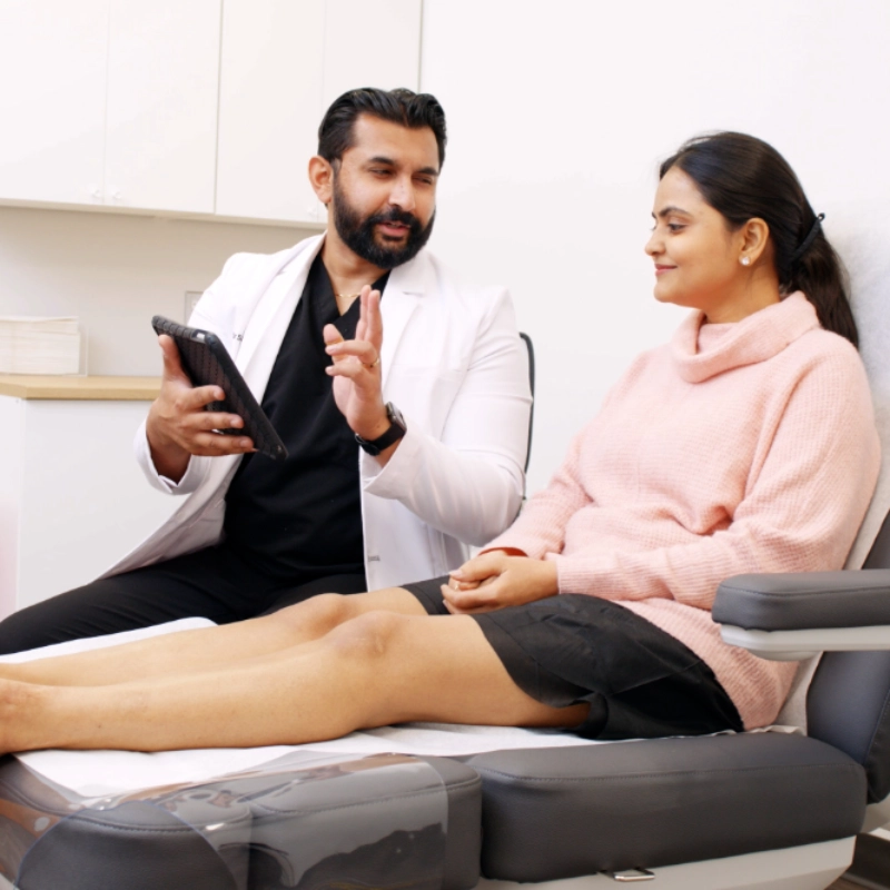 A friendly doctor in a white lab coat shows information about a vein treatment process on a tablet to a seated, smiling patient in a modern medical office setting.