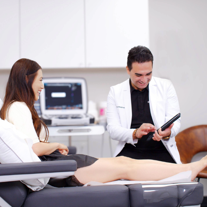 A friendly doctor in a white lab coat shows information about a vein treatment process on a tablet to a seated, smiling patient in a modern medical office setting.
