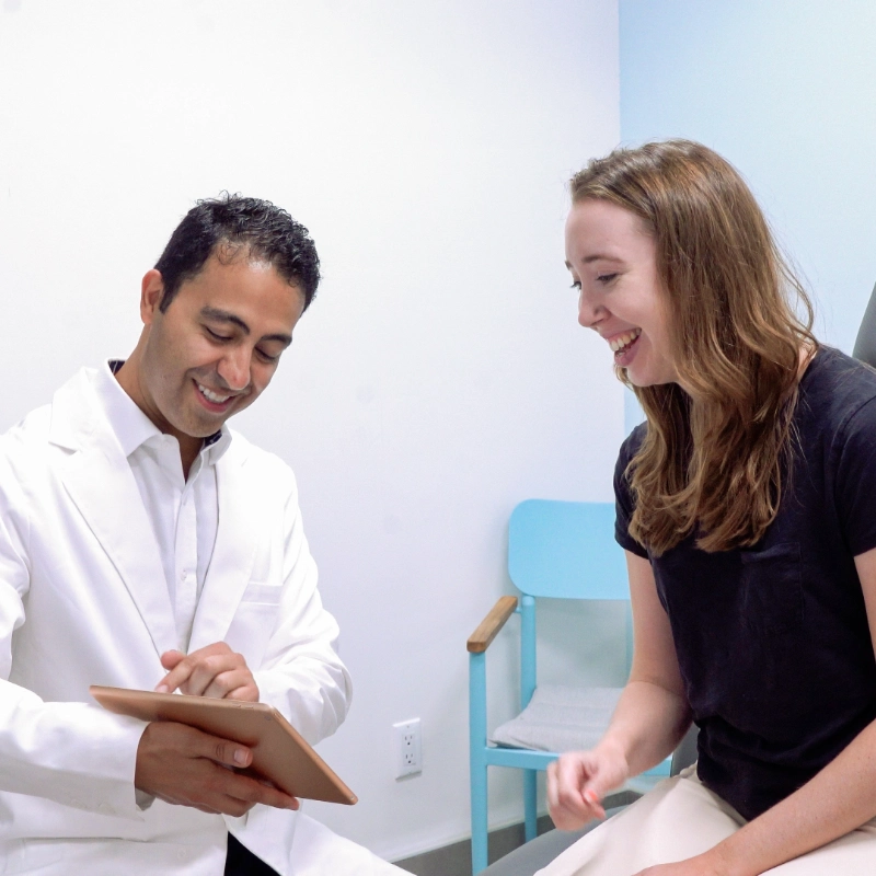 Dr. Kamran Saraf is consulting with a female patient in a clinical examination room. The doctor is showing the patient information on a tablet, and both are smiling during the positive interaction.