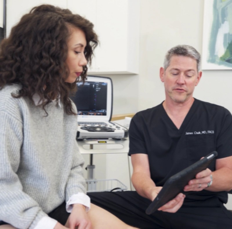 A friendly doctor in a white lab coat shows information about a vein treatment process on a tablet to a seated, smiling patient in a modern medical office setting.