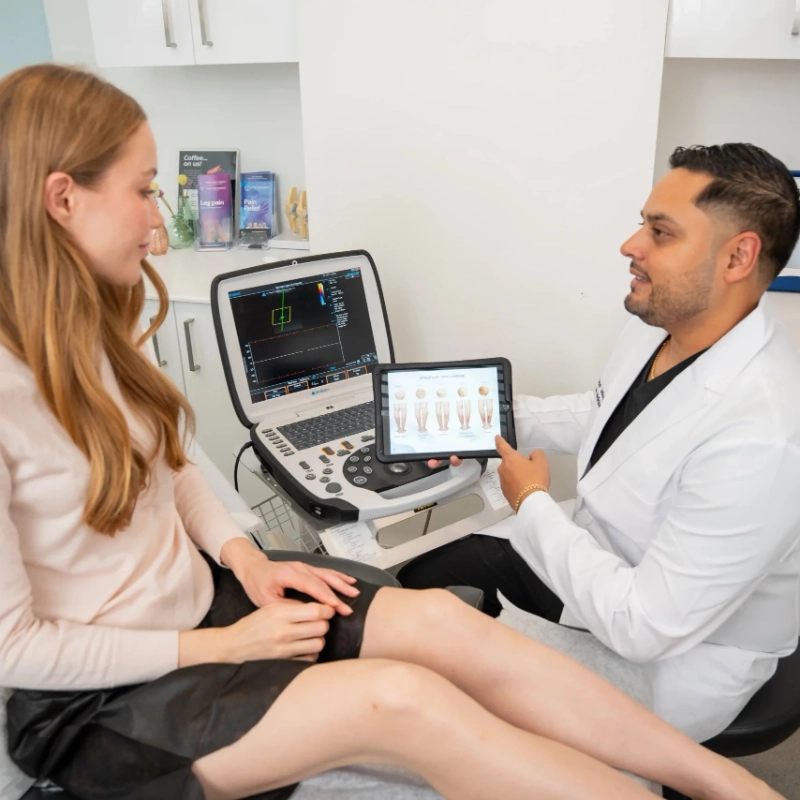 A friendly doctor in a white lab coat shows information about a vein treatment process on a tablet to a seated, smiling patient in a modern medical office setting.