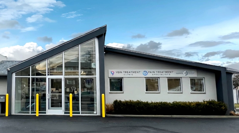 Exterior view of a medical facility featuring the "Vein Treatment Clinic" and "Pain Treatment Specialists" signs. The building has a modern grey facade with a large slanted glass entrance, located next to a brick building under a blue sky.