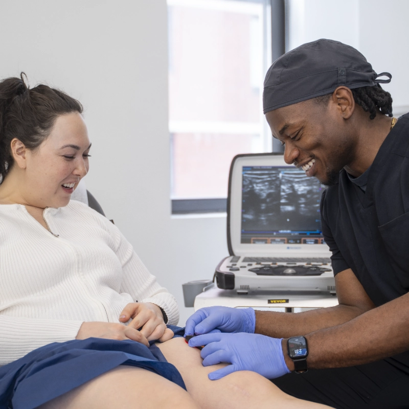 Dr. Baron Ekeledo performs a Duplex Ultrasound examination on a patient's lower extremity in a clinical setting, with the patient appearing engaged and comfortable during the interaction. The Ultrasound Machine is visible in the background with imaging results displayed on its monitor, demonstrating a professional vascular assessment procedure.