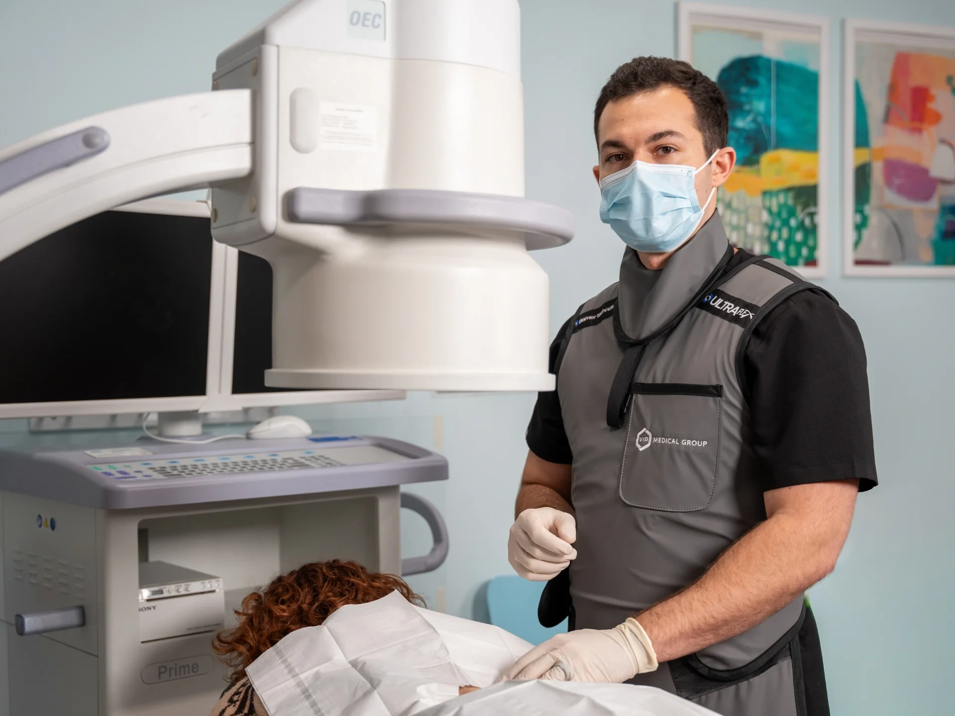 Dr. Kyle Mele, wearing a lead apron and surgical mask, stands beside a patient during a medical procedure. A C-arm fluoroscopy machine is positioned over the patient to provide real-time imaging for the intervention.