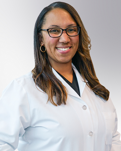 Professional headshot of Dr. Tamara Green wearing a white lab coat over dark scrubs and glasses, smiling confidently against a black background. This official clinical portrait represents a healthcare provider ready for patient consultations and medical practice.