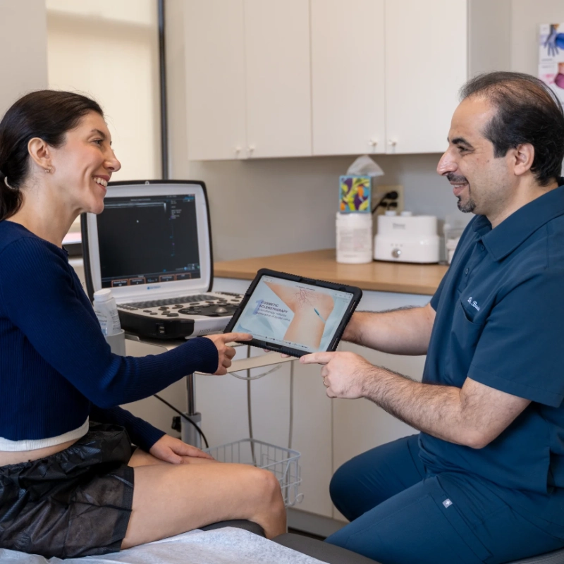 Dr. Mohsen Basiri consults with a female patient in a clinical office setting, utilizing a tablet device to review diagnostic imaging or educational content related to venous conditions. The provider demonstrates and explains clinical findings while the patient sits attentively. The background displays a medical office equipped with an ultrasound machine, diagnostic software on a laptop, and various medical supplies.