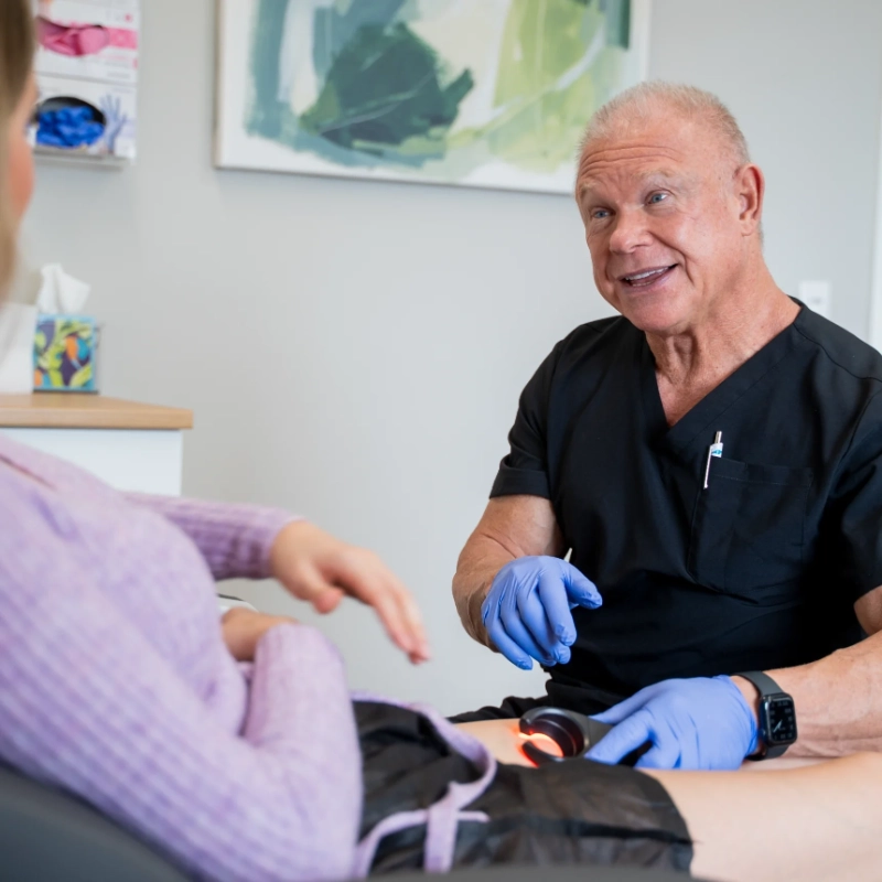 Dr. David Stone engages with a patient during a clinical examination in a medical office setting. The provider, wearing black scrubs and blue gloves, performs a lower extremity physical examination with a professional and approachable demeanor. The clean clinical environment with basic furnishings is visible in the background.