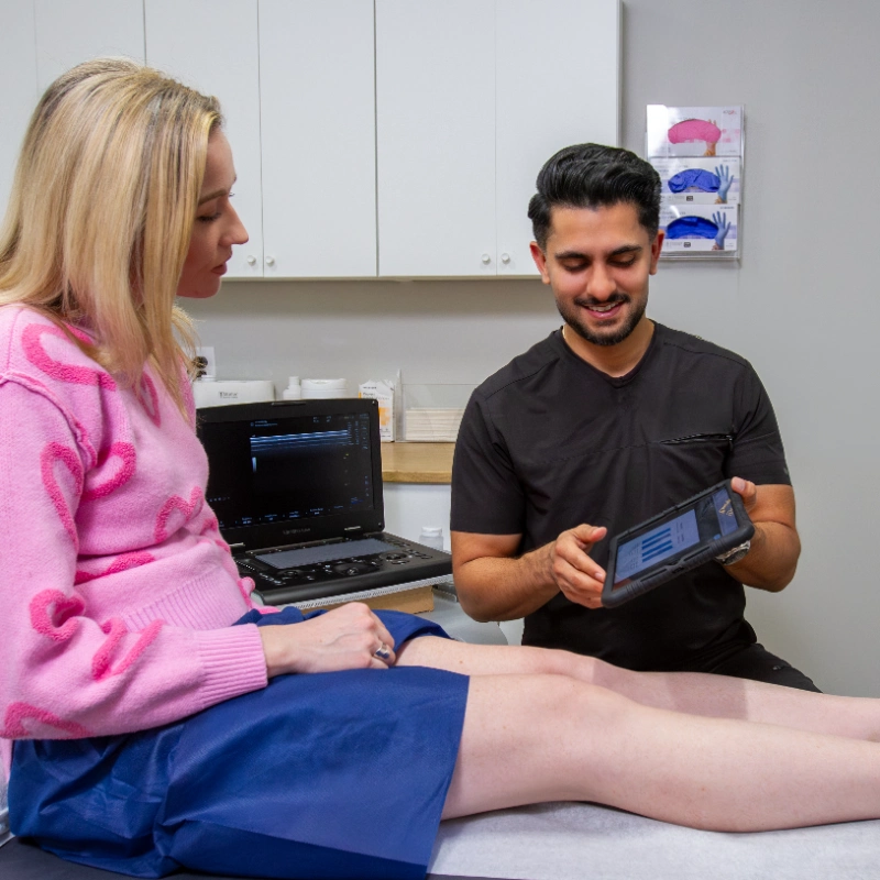 Dr. Akash Mehta conducts a Duplex Ultrasound examination of the patient's lower leg and foot to assess for Venous Reflux and Chronic Venous Insufficiency. The provider uses a portable Ultrasound Machine while an Ultrasound Machine with imaging software is visible on the desk in the background of this modern clinical examination room.