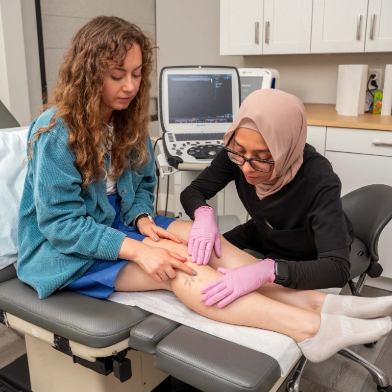 Dr. Anee Khan performs a physical examination of a patient's lower leg and calf area for venous assessment while the patient sits on an examination table in a clinical setting. An Ultrasound Machine is visible in the background, suggesting preparation for Duplex Ultrasound evaluation to assess for Varicose Veins or Chronic Venous Insufficiency.