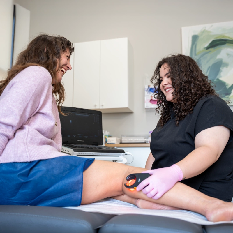 Dr. Hanen Fernandez, MD performs a Duplex Ultrasound examination on a patient's leg using an Ultrasound Machine with an Ultrasound Probe while wearing pink gloves. The patient is seated on an examination table in a clinical treatment room, with ultrasound imaging displayed on a laptop in the background. This diagnostic procedure is used to evaluate venous conditions including Varicose Veins, Spider Veins, Chronic Venous Insufficiency, and Venous Reflux