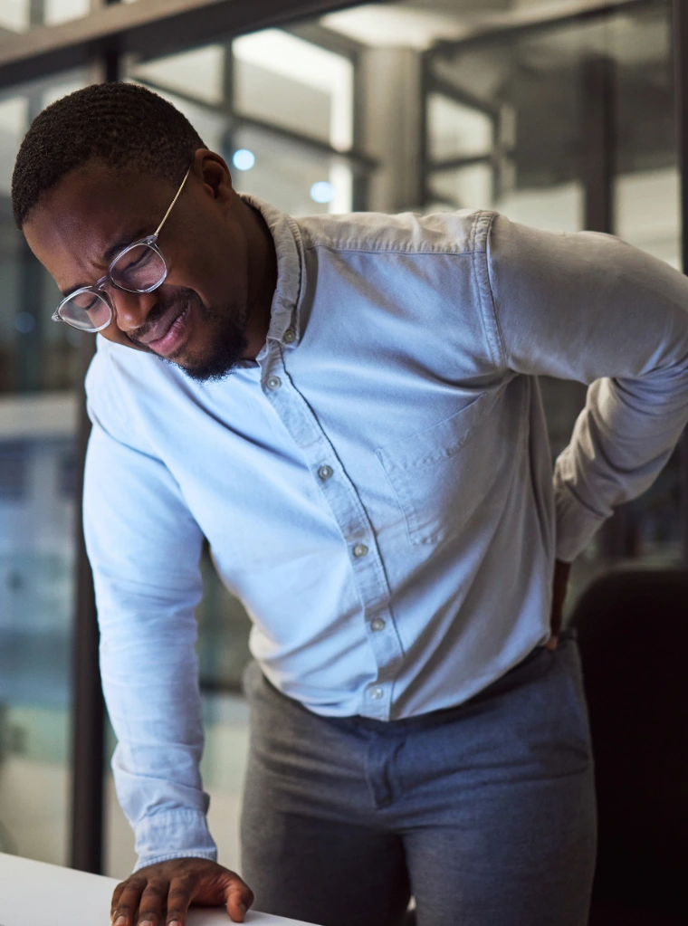 Back_pain_condition A man wearing glasses and a light blue button-down shirt leaning against a white desk, wincing in discomfort while clutching his lower back with one hand.