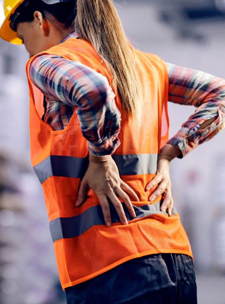 Spine_pain_condition_optimized A female industrial worker wearing a yellow hard hat and bright orange high-visibility safety vest, standing on a job site with both hands pressed against her lower back in discomfort.